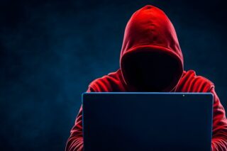 Eine mysteriöse Gestalt in einem roten Kapuzenpullover sitzt vor einem Laptop, Cybercrime-Foren