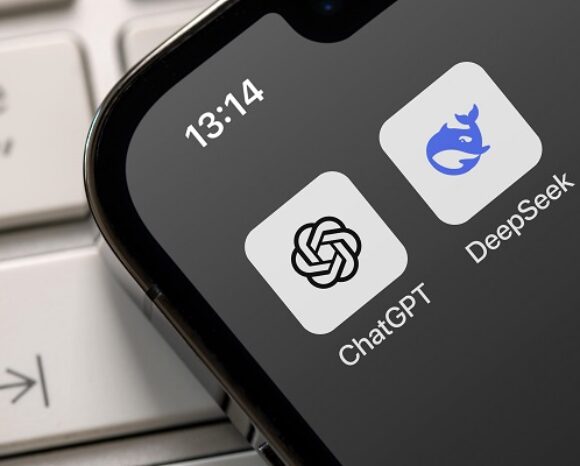 Nahaufnahme eines Smartphone-Bildschirms mit zwei App-Symbolen: ChatGPT mit einem schwarzen Wirbel-Logo und DeepSeek mit einem blauen Wal-Logo. Die angezeigte Uhrzeit ist 13:14 Uhr. Das Telefon liegt auf einer Laptop-Tastatur.