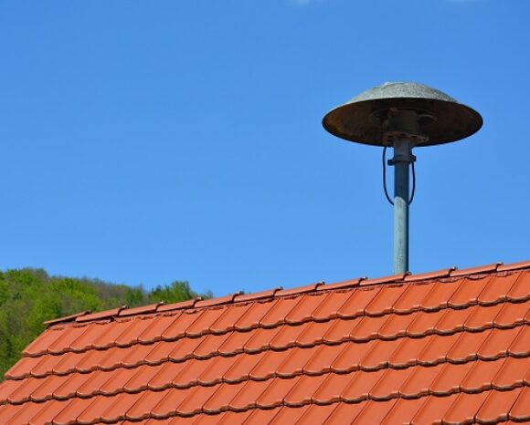 Ein rotes Ziegeldach mit einer historischen Sirene auf einem Metallmast. Der Himmel ist klar und blau, im Hintergrund ist ein baumbedeckter Hügel zu sehen.