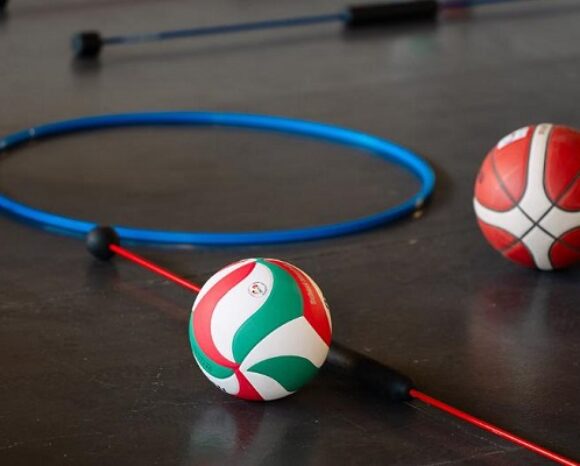 Zwei Bälle und ein blauer Hula-Hoop-Reifen liegen auf einem Hallenboden; im Vordergrund ein Volleyball mit grün-weiß-rotem Muster, daneben ein roter Gymnastikstab, im Hintergrund ein Basketball.