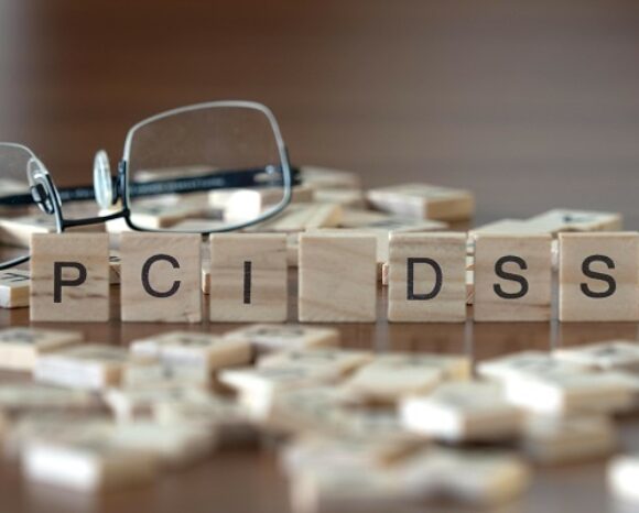 Das Akronym "PCI DSS" für Payment Card Industry Data Security Standard dargestellt durch hölzerne Buchstabensteine ​​auf einem Holztisch mit einer Brille und einem Buch