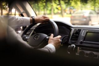 Hände am Lenkrad in einem Pkw; Sicht aus dem Innenraum auf Armaturenbrett, Infotainment und vorausfahrenden Verkehr bei Tageslicht; Fokus auf Fahrsicherheit und Straßenverkehr.