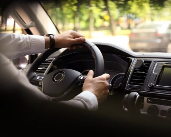 Hände am Lenkrad in einem Pkw; Sicht aus dem Innenraum auf Armaturenbrett, Infotainment und vorausfahrenden Verkehr bei Tageslicht; Fokus auf Fahrsicherheit und Straßenverkehr.