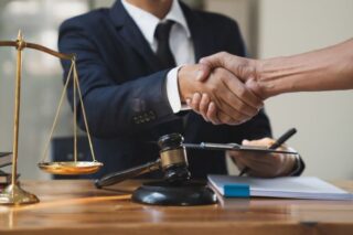 Anwalt und Mandant geben sich die Hand über einem Schreibtisch mit Justizwaage und Richterhammer, Abschluss einer Rechtsberatung oder Einigung.