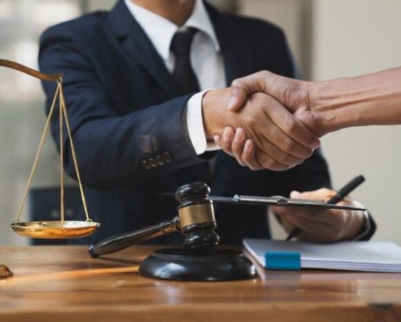 Anwalt und Mandant geben sich die Hand über einem Schreibtisch mit Justizwaage und Richterhammer, Abschluss einer Rechtsberatung oder Einigung.