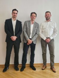 Drei Kollegen in Businesskleidung stehen nebeneinander vor heller Wand im Büro; formelles Teamfoto, frontal und freundlich, Business-Porträt auf Parkettboden.